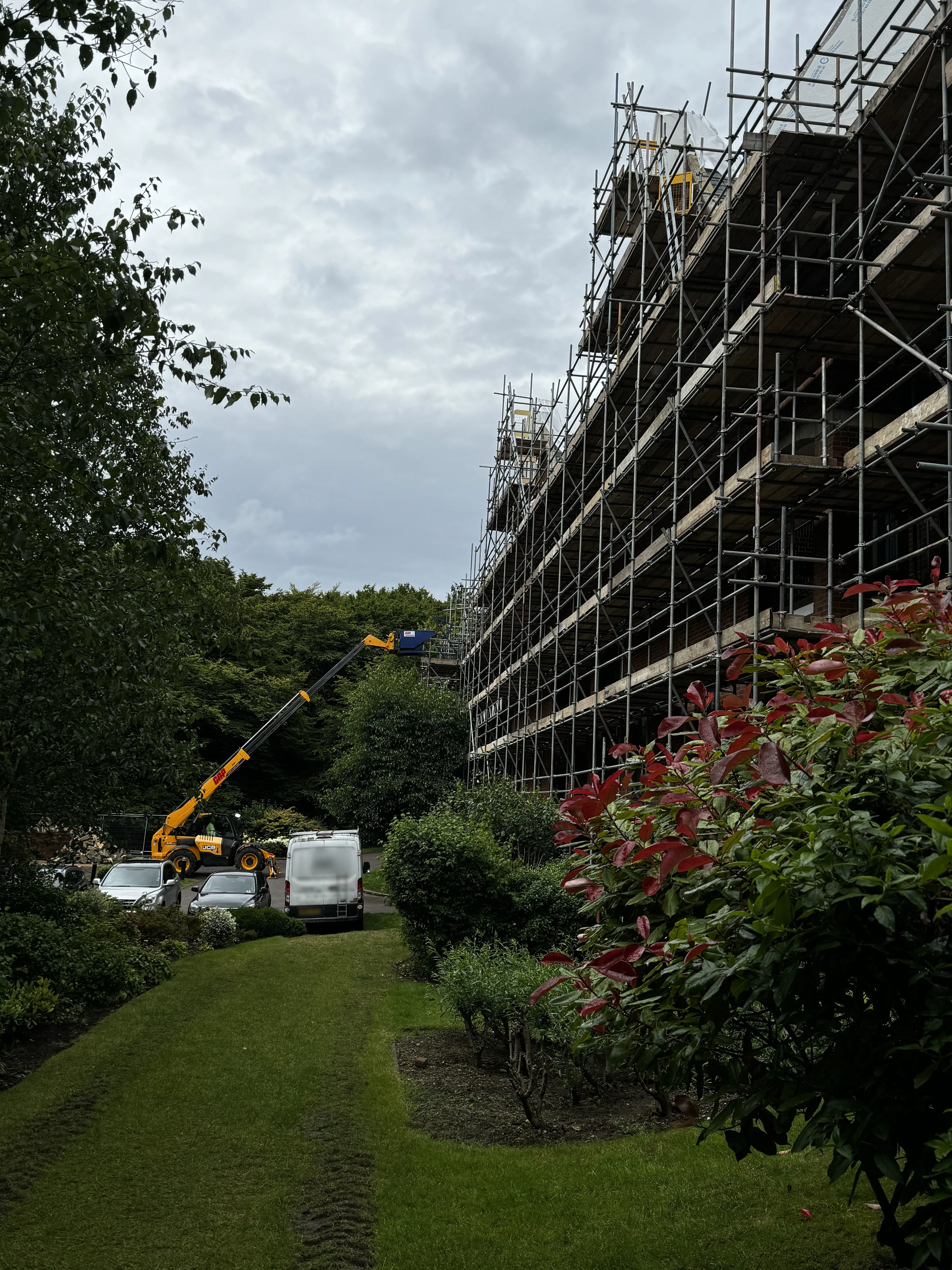 A large Victorian building fully scaffolded for reinstatement, seen across the lawn with a telehandler and site vehicles