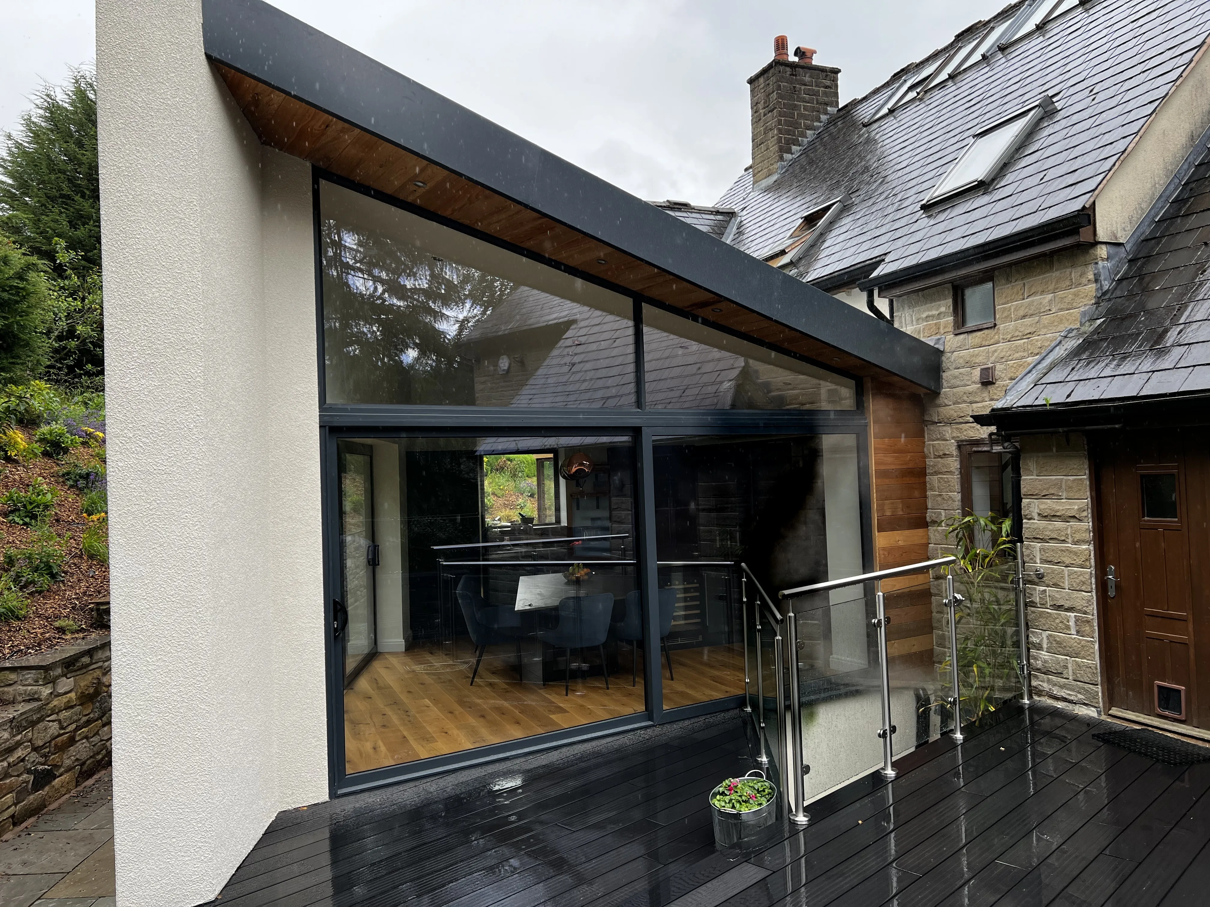 The completed extension on a wet day: white render fin, cedar soffit and full-height glazing against the stone house, with a composite deck