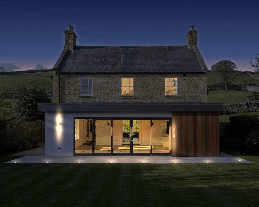 Illustration: a finished single-storey rear extension on a stone house at dusk, lit from inside: white render, cedar cladding, sliding doors and a porcelain patio.